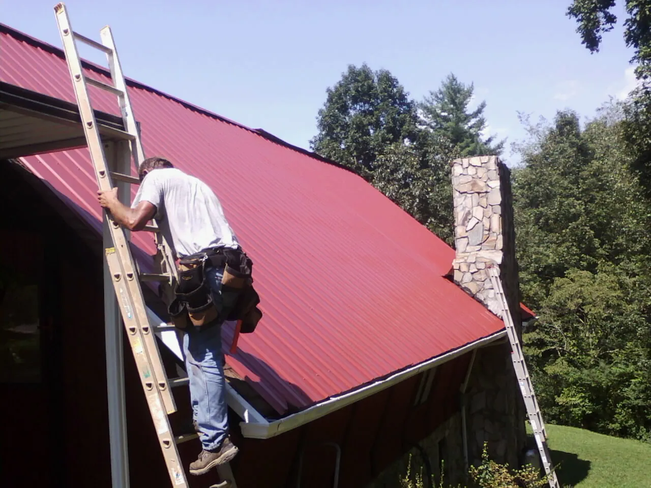 Roofer on a ladder beside a red metal roof and stone chimney — Soffit & Fascia Repair by Roofing Brothers in Swain